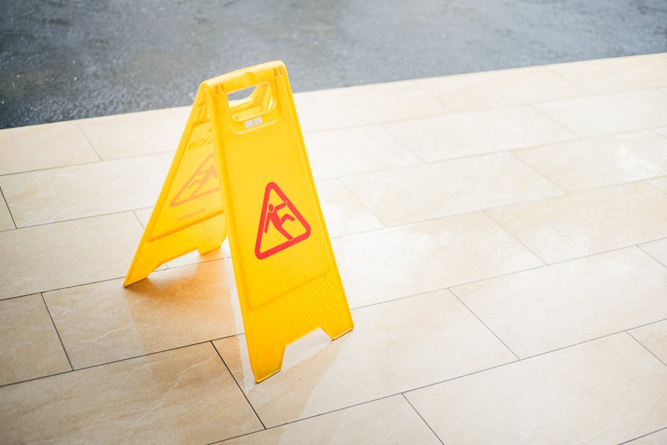 A yellow A-frame wet floor warning sign with red triangle symbols and a black exclamation mark and person slipping, positioned on a beige tiled floor. The sign warns of a potential hazard on the surfa