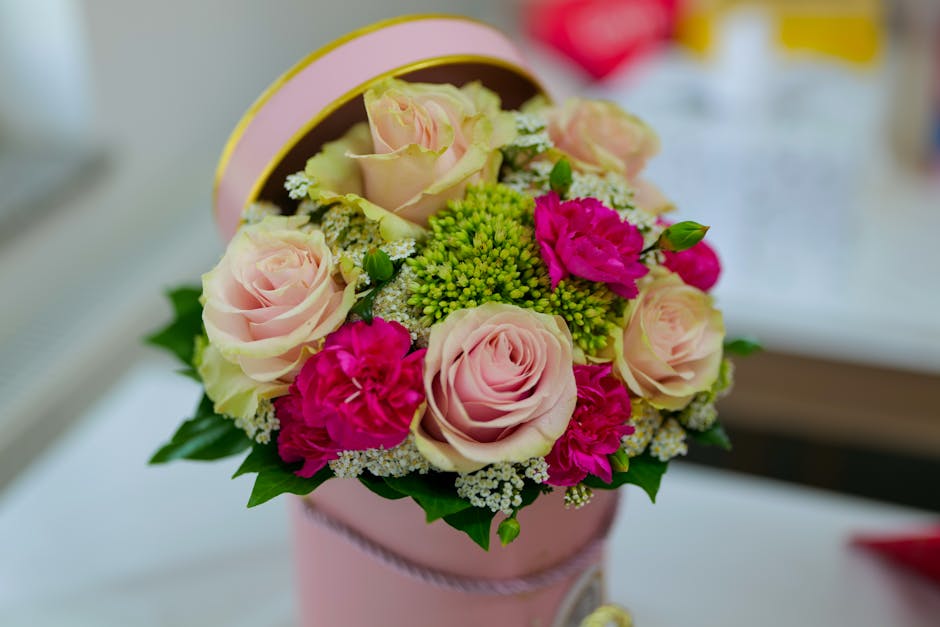 A round floral arrangement in a light pink cylindrical box featuring pale pink roses, bright pink carnations, small white daisies, and vibrant green filler flowers, all arranged with lush green foliag